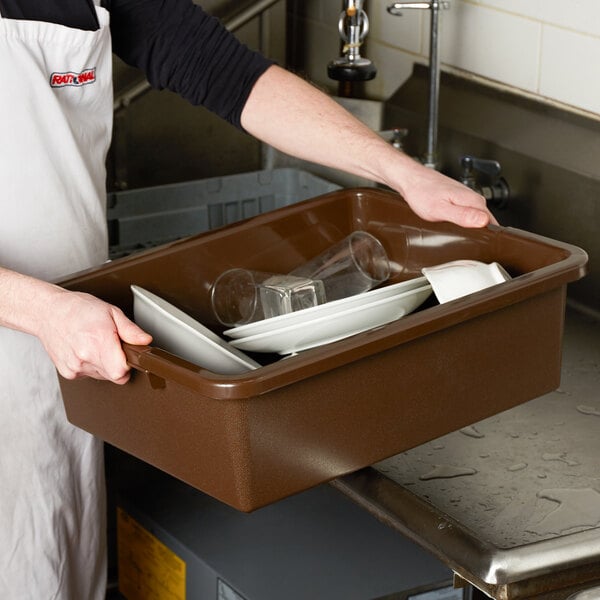 A man in an apron holding a brown Tablecraft bus tub on a counter.