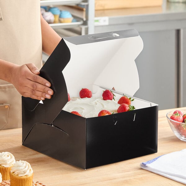 A black 10 x 10 x 5 inch bakery box containing a cake topped with strawberries.