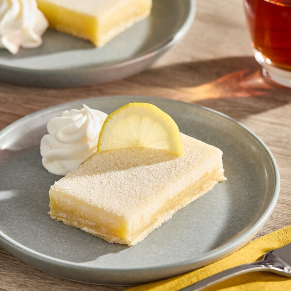 A rectangular lemon bar square garnished with a lemon slice and a dollop of whipped cream on a gray plate.