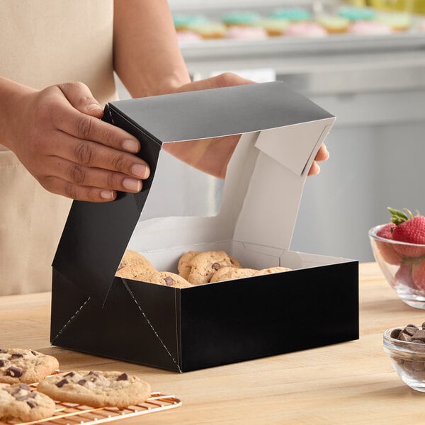 A black auto-popup bakery box filled with cookies, being opened on a kitchen counter.