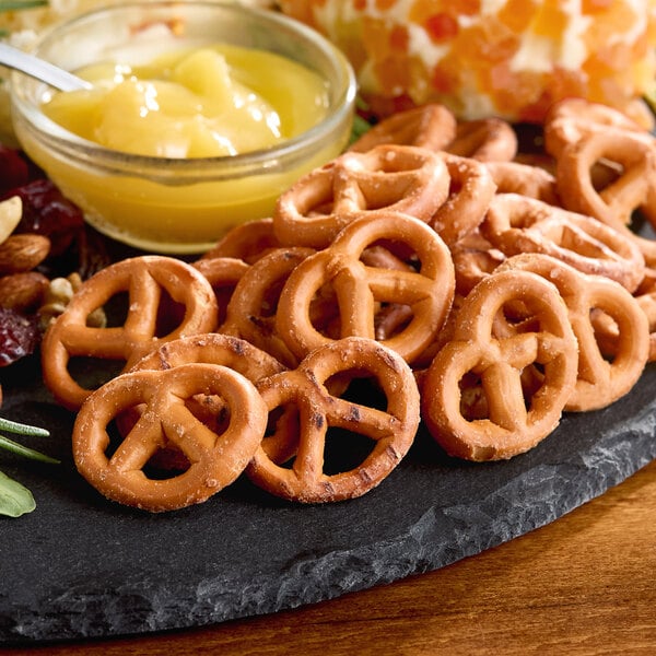 A pile of small, salted gluten-free pretzels on a black serving tray with a bowl of yellow dip in the background.