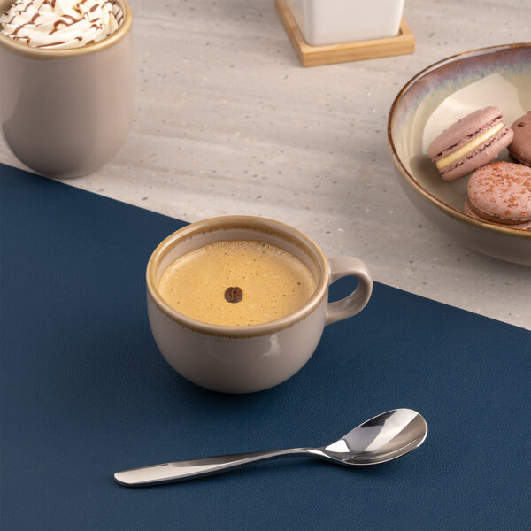 A beige porcelain tea cup filled with a hot beverage, placed on a blue surface with a spoon nearby.