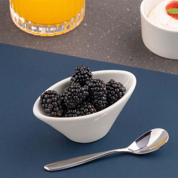 A small white bowl filled with blackberries is placed next to a metal spoon on a blue surface, with a glass of orange juice and a dessert partially visible in the background.