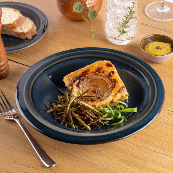A blue round porcelain coupe deep plate with a slice of savory tart and garnishes, set on a wooden table.