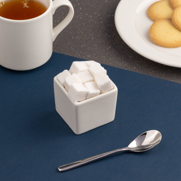 A small ivory square porcelain bowl filled with sugar cubes, placed next to a spoon.