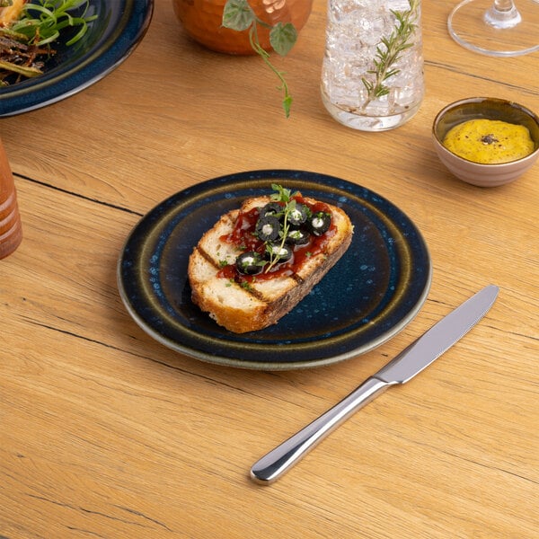 A blue round porcelain coupe flat plate with a slice of bread topped with garnish, placed on a wooden table.