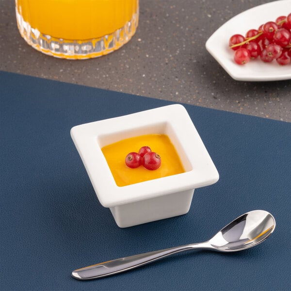 A small ivory square porcelain bowl with a wide rim, filled with a yellow dessert and garnished with red berries.