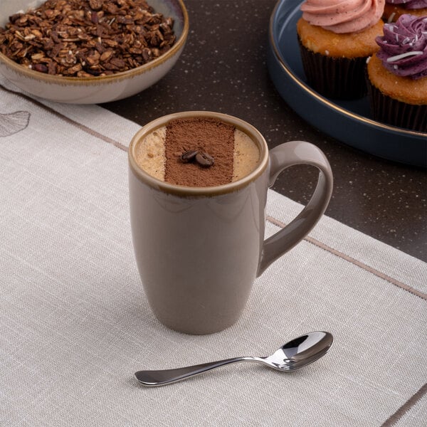 A beige tapered porcelain mug filled with coffee, placed on a table with a spoon nearby.