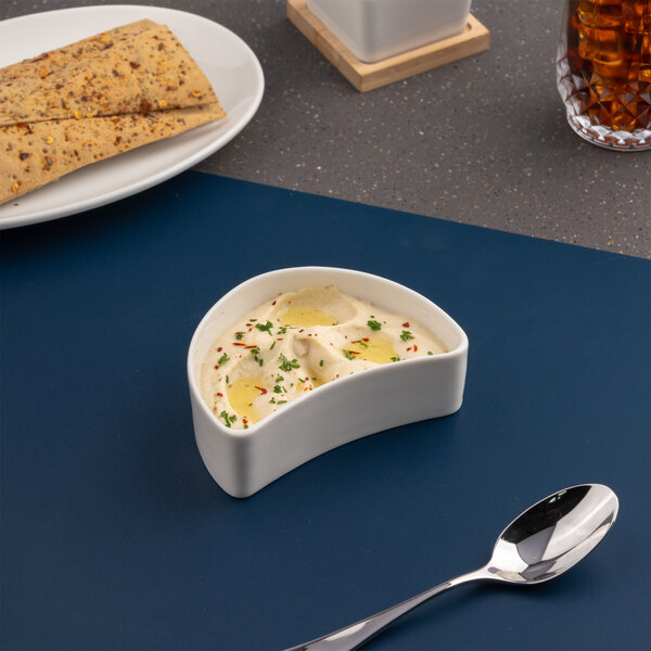 A small ivory half round porcelain coupe bowl filled with a creamy dip, placed on a blue surface.