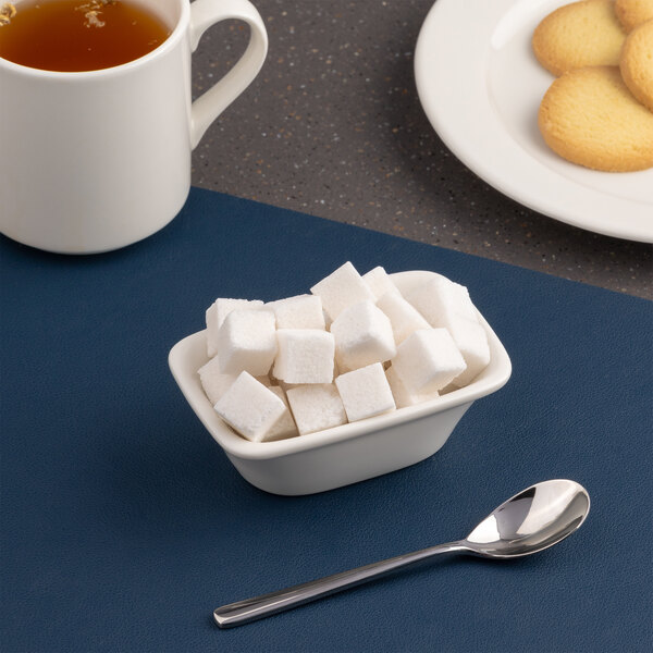 A small ivory rectangular porcelain bowl filled with sugar cubes, placed next to a spoon, a cup of tea, and a plate of cookies.