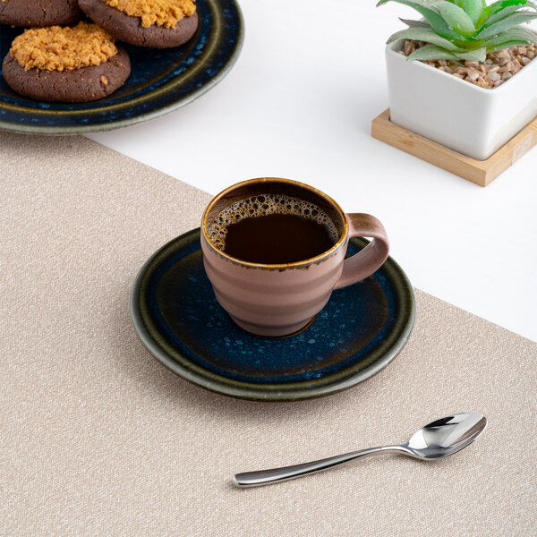 A blue porcelain espresso cup with a matching saucer, filled with coffee and placed on a beige placemat next to a spoon.