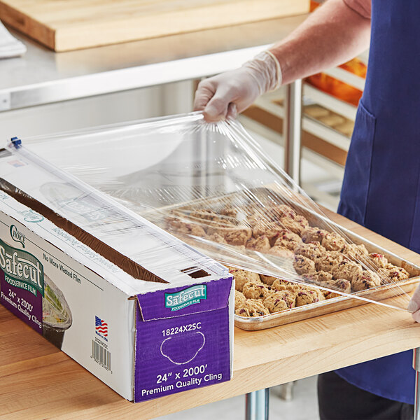 A box of Choice Safecut 24-inch by 2000-foot premium foodservice film with a slide cutter being used to cover a tray of food.