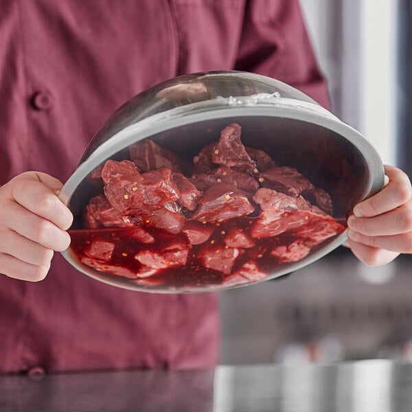 A person holding a metal bowl filled with marinated raw meat pieces.