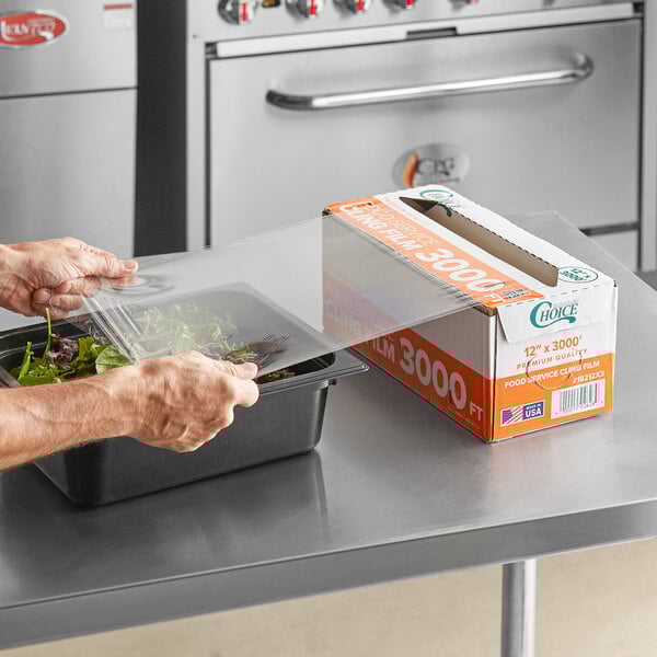 A box of Choice 12-inch by 3000-foot foodservice film with a serrated cutter being used to cover a food container.