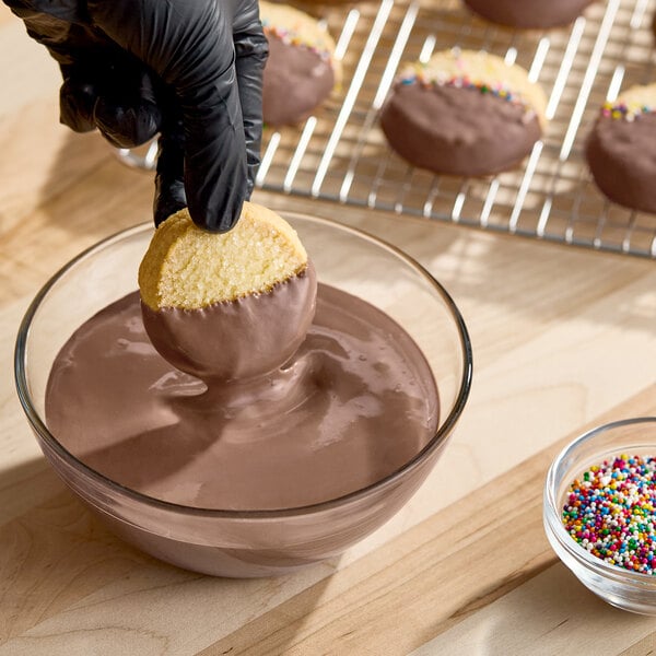 A bowl of melted milk chocolate compound coating being used to dip cookies, with finished chocolate-coated cookies and sprinkles nearby.