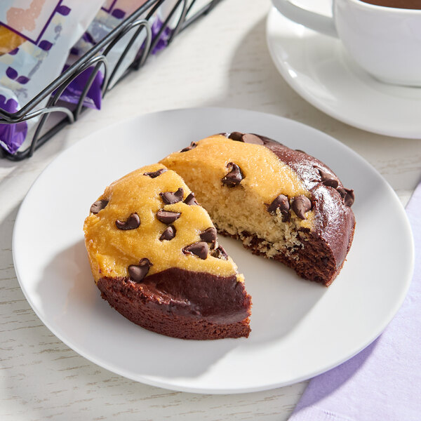A round, individually wrapped vegan tuxedo cake with chocolate and vanilla sections topped with chocolate chips, served on a white plate.