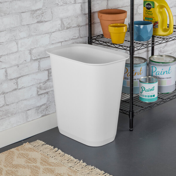 A white rectangular plastic wastebasket placed on the floor near a shelving unit.