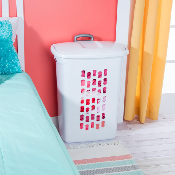 A white rectangular laundry hamper with a flip-top lid and wheels, placed next to a bed.