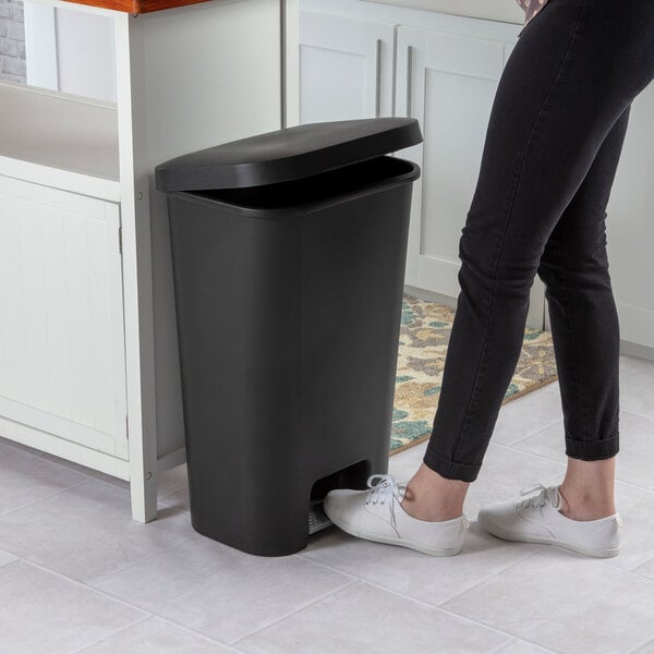 A black rectangular step-on wastebasket with a partially open lid, placed in a kitchen setting.