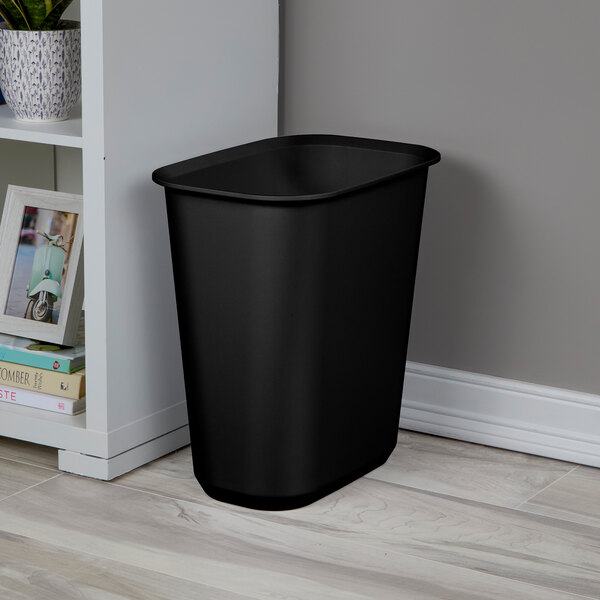 A black rectangular plastic wastebasket placed on a floor next to a white shelf.