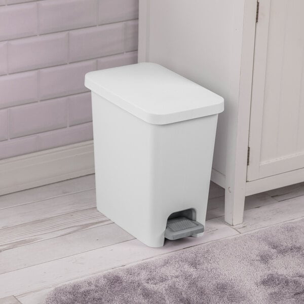 A white rectangular step-on wastebasket with a foot pedal, placed next to a cabinet on a bathroom floor.