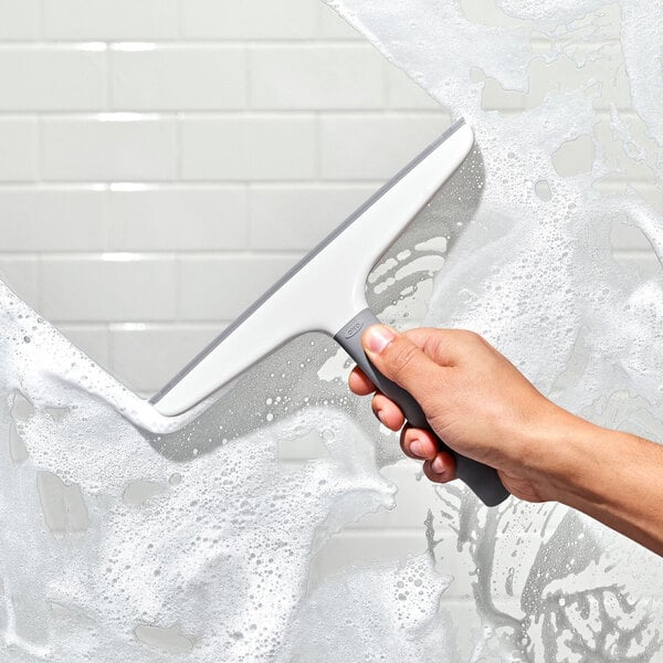A white and gray plastic squeegee with a hook being used to clean soapy water from a tiled surface.