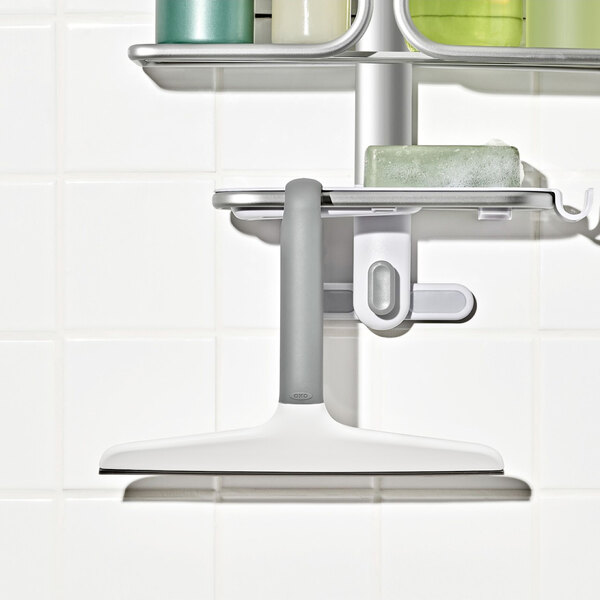 A white and gray plastic squeegee with a hook, hanging on a shower caddy against a tiled wall.
