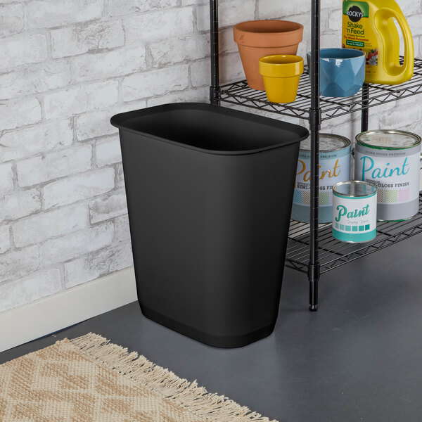 A black rectangular 9-gallon wastebasket placed on the floor next to a shelving unit.