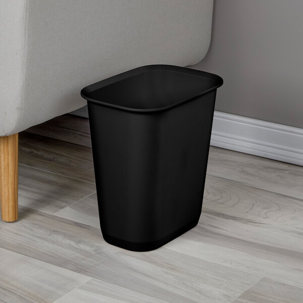 A black rectangular plastic wastebasket placed on a wooden floor next to a couch.