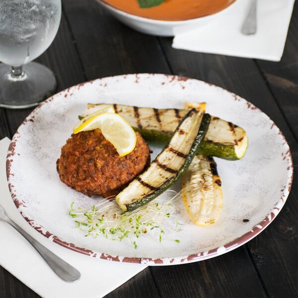 A white, irregular round melamine coupe plate with a rustic brown rim, holding a serving of grilled vegetables and a patty garnished with a lemon slice.