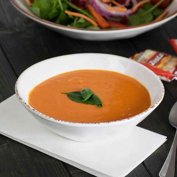A white melamine bowl filled with tomato soup, garnished with basil, placed on a white napkin.