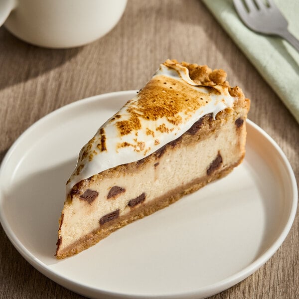 A slice of s'mores cheesecake with a graham cracker crust, chocolate chunks, and toasted marshmallow topping on a white plate.