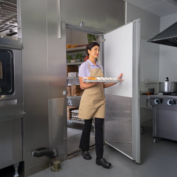 A person carrying a tray of food is walking out of a Kolpak custom walk-in cooler or freezer unit in a commercial kitchen.