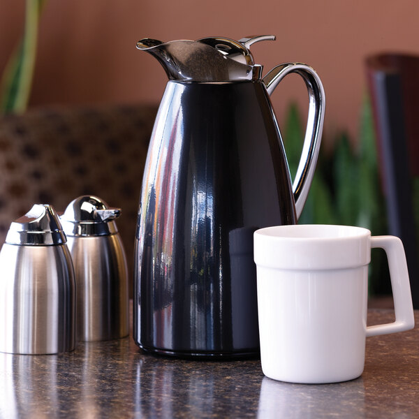 A black powder-coated stainless steel insulated coffee carafe with a push-button lid, shown next to a white mug and two small metal containers.