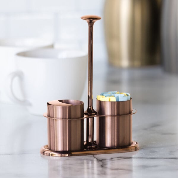 A rose gold stainless steel sugar stick holder and creamer set with a central handle.