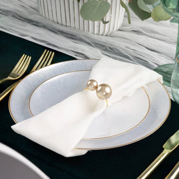 A round white plastic plate with a gold band, set on a table with gold utensils and a white napkin.