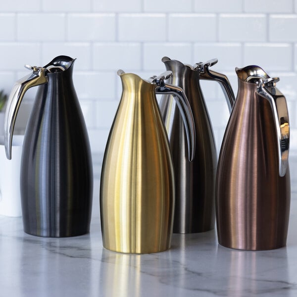 A group of four stainless steel insulated coffee carafe servers in different metallic finishes, including dark tungsten, gold, bronze, and black, displayed on a countertop.