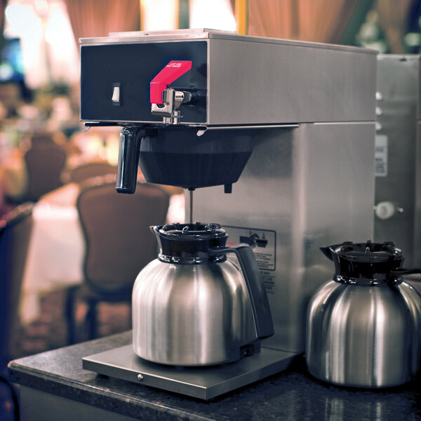 A stainless steel coffee server with a black brew-thru lid placed under a commercial coffee maker.