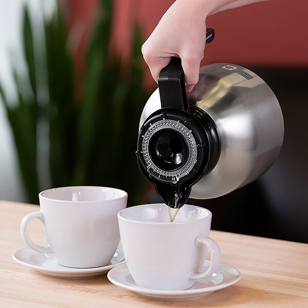 A stainless steel coffee server with a black handle and orange clock lid pouring coffee into a white cup on a saucer next to another cup.