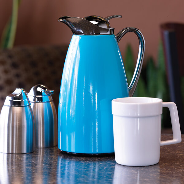 A blue powder-coated stainless steel insulated coffee carafe with a push-button lid, shown alongside a white mug and two small metal containers.