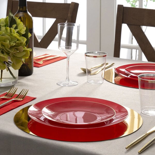 A red round plastic plate with a gold band, set on a dining table with gold utensils and glassware.