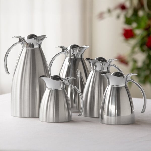 A brushed stainless steel insulated carafe with a flip-top lid, shown among other similar carafes.