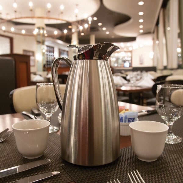 A brushed stainless steel insulated coffee server with a handle and spout, placed on a restaurant table setting.