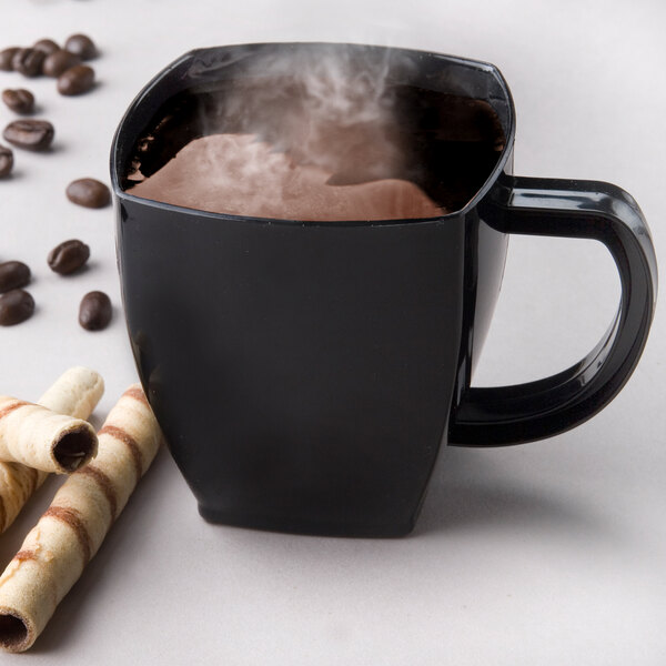 A black square-shaped disposable plastic coffee mug filled with a hot beverage, shown with coffee beans and wafer rolls nearby.