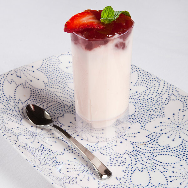 A clear, triangular polystyrene tumbler filled with a dessert, garnished with a strawberry slice and mint, placed on a patterned plate with a spoon.