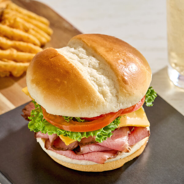 A sandwich made with a sliced milk bun, filled with lettuce, tomato, cheese, and sliced meat, served with waffle fries in the background.
