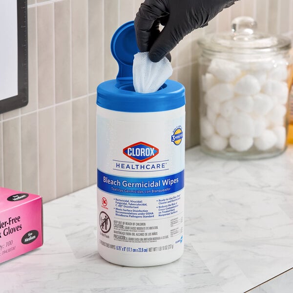 A container of Clorox Healthcare Bleach Germicidal Wipes with a blue lid, being used by a person wearing a black glove.