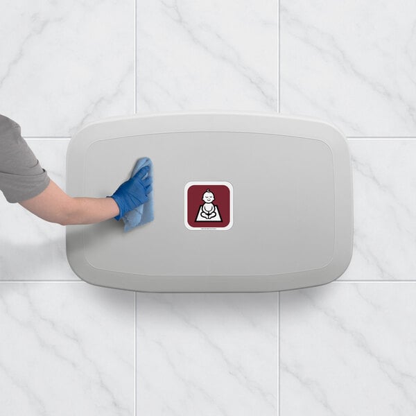 A gray horizontal surface-mounted baby changing station being cleaned by a person wearing a blue glove.