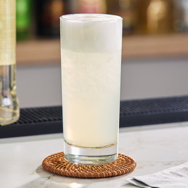 A clear, straight-sided 10.5 oz. Collins glass filled with a light-colored beverage, placed on a woven coaster.