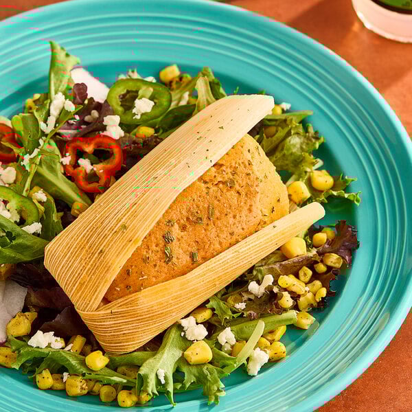 A red chile and pork tamale served on a bed of mixed greens, corn, and sliced peppers on a turquoise plate.